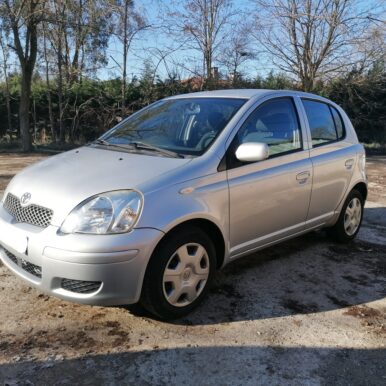 Toyota Yaris argentée 5 portes stationnée sur un terrain extérieur ensoleillé avec arbres en arrière-plan.